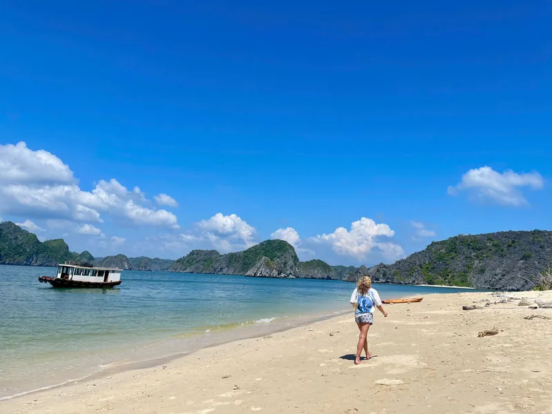 A woman walks along a quiet sandy beach with a boat anchored offshore amid limestone islands in Lan Ha Bay.