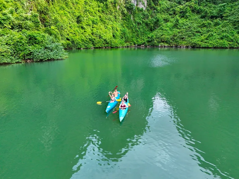 Two kayakers paddle turquoise green water beneath forested limestone cliffs in Lan Ha Bay, Cat Ba.