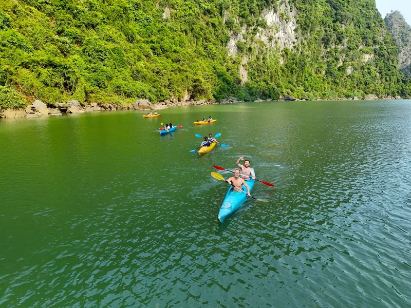 Kayakers paddle colorful boats across calm green water beneath limestone karsts in Lan Ha Bay.