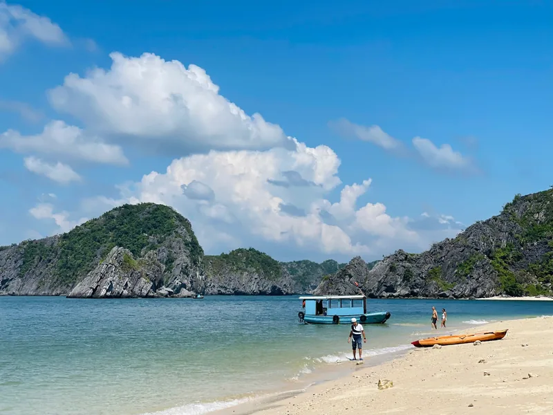 A sandy beach in Lan Ha Bay with limestone karsts, a small boat and kayak, and people wading in clear water.