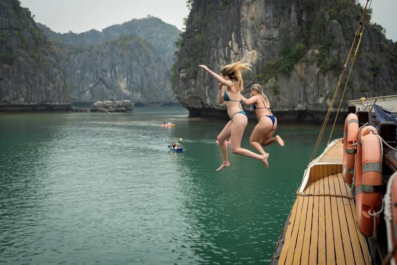 Two women in bikinis leap off a cruise boat deck into the emerald waters of Lan Ha Bay, surrounded by limestone karsts.