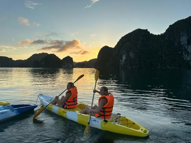 Two kayakers in orange life vests paddle a yellow kayak at sunset among the limestone karsts of Lan Ha Bay.