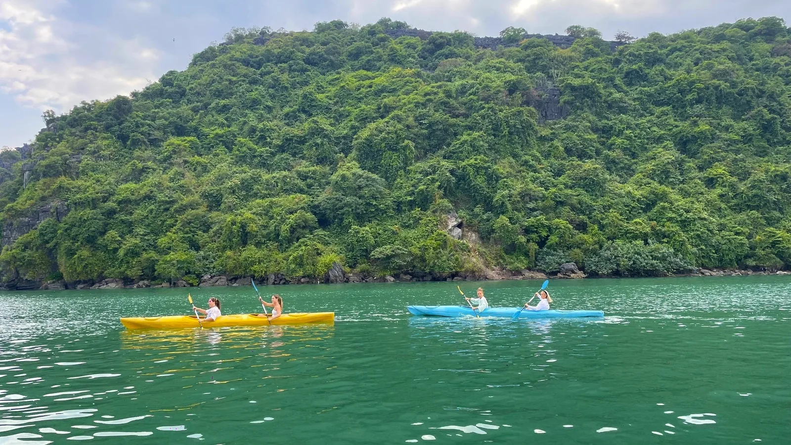 Kayakers paddle yellow and blue kayaks across emerald waters beneath a forested limestone cliff in Lan Ha Bay.