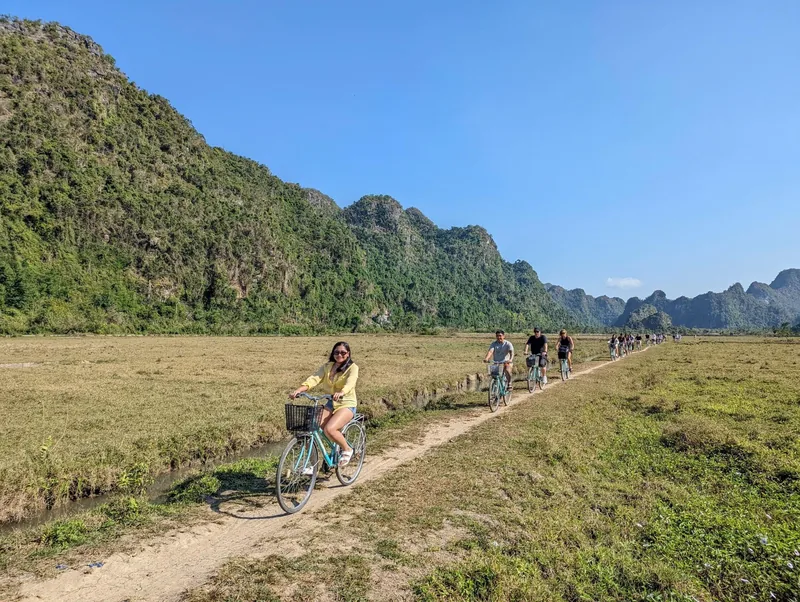 Cyclists ride a narrow dirt path through green fields flanked by limestone karsts in Viet Hai village, Cat Ba.