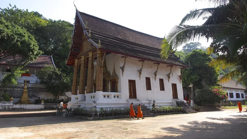 Monks in orange robes walk past a whitewashed temple hall with a steep tiered roof in Luang Prabang.