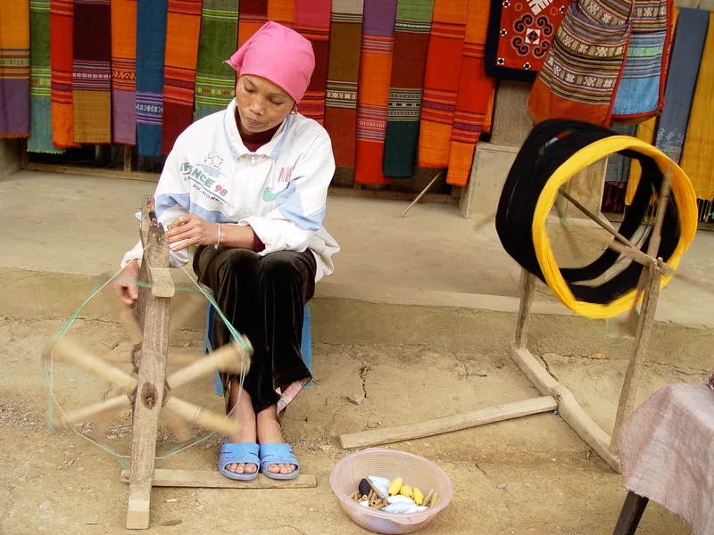 A woman in Mai Chau spins thread on a wooden wheel beside colorful woven fabrics on display.