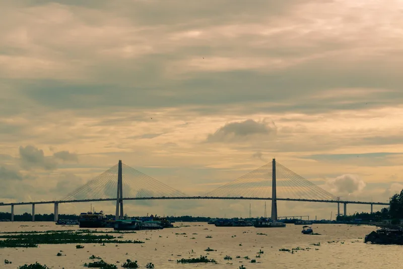 A cable-stayed bridge spans the wide Mekong River under a golden sunset sky, with boats scattered on the water below.