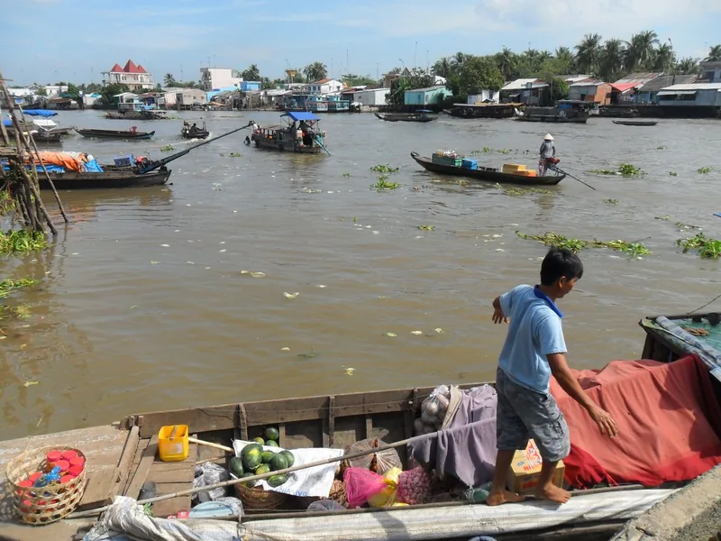 A man stands on a fruit-laden boat as small vessels drift along a Mekong Delta river lined with houses.