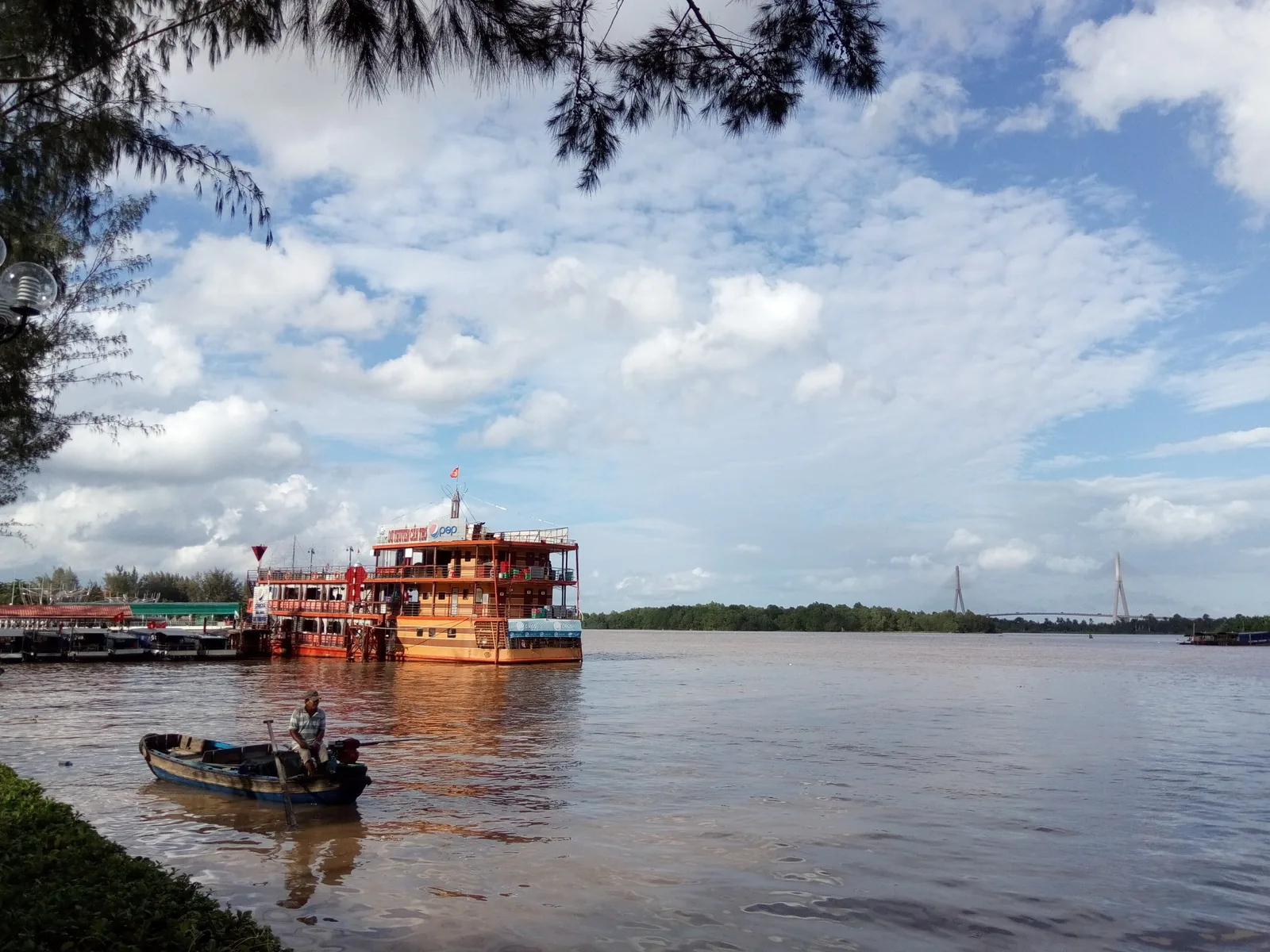 A wooden tour boat and small fishing craft float on the Bassac River near Ninh Kieu in Can Tho, with a cable bridge in the.