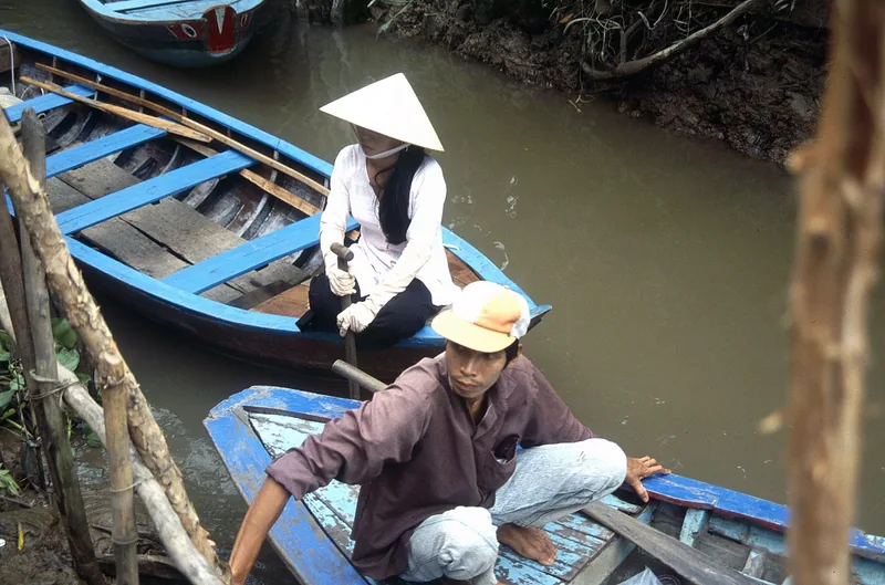A woman in a conical hat and a man paddle small wooden boats along a muddy Mekong Delta canal.