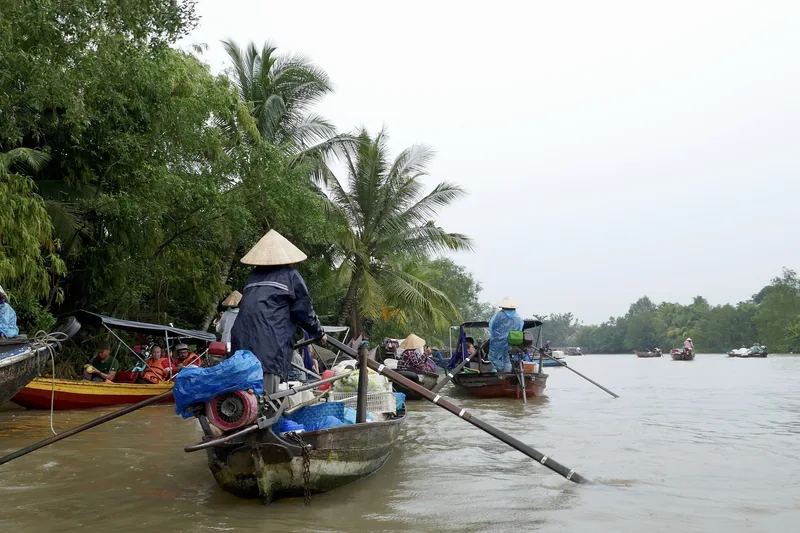 Vendors in conical hats row wooden boats along a river at a floating market in the Mekong Delta.