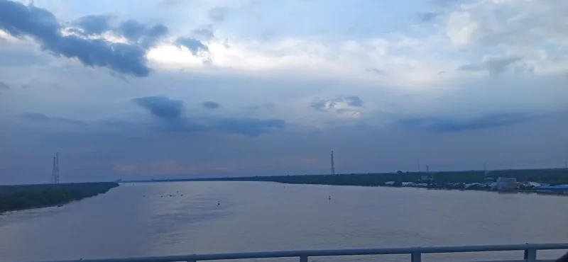 A wide, muddy river flows under a cloudy evening sky, viewed from the Ham Luong Bridge in the Mekong Delta.