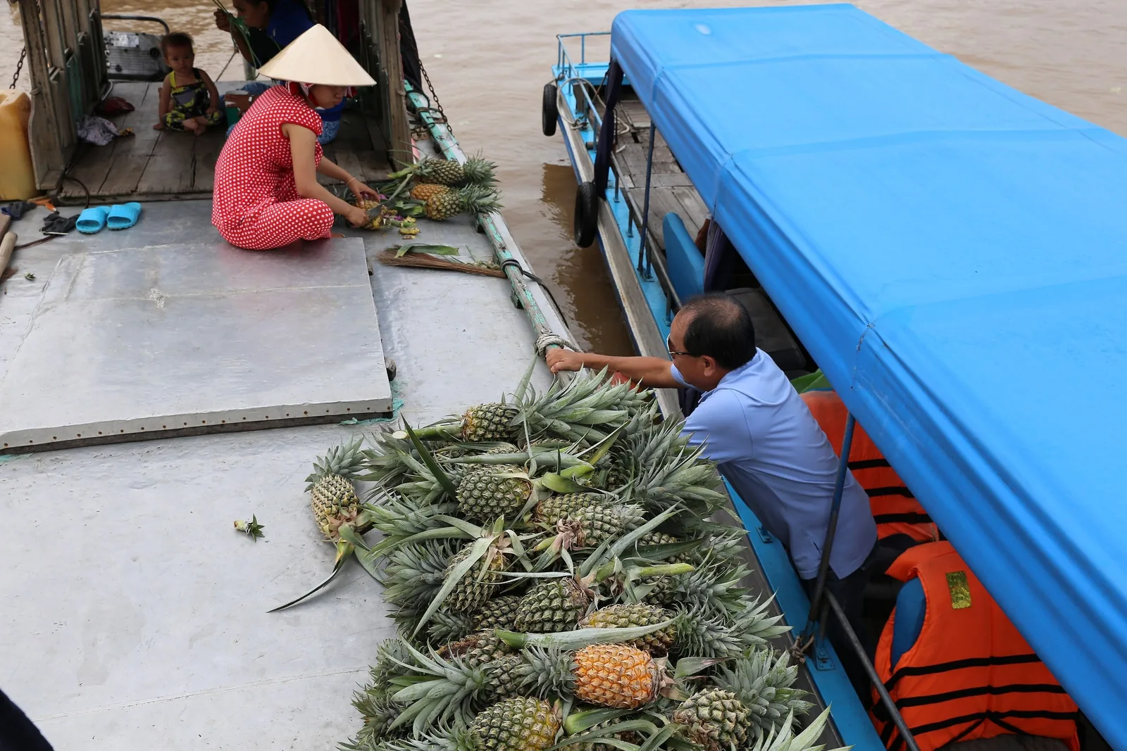 A vendor in a conical hat prepares pineapples on a river boat while a tourist reaches for the produce near Can Tho.