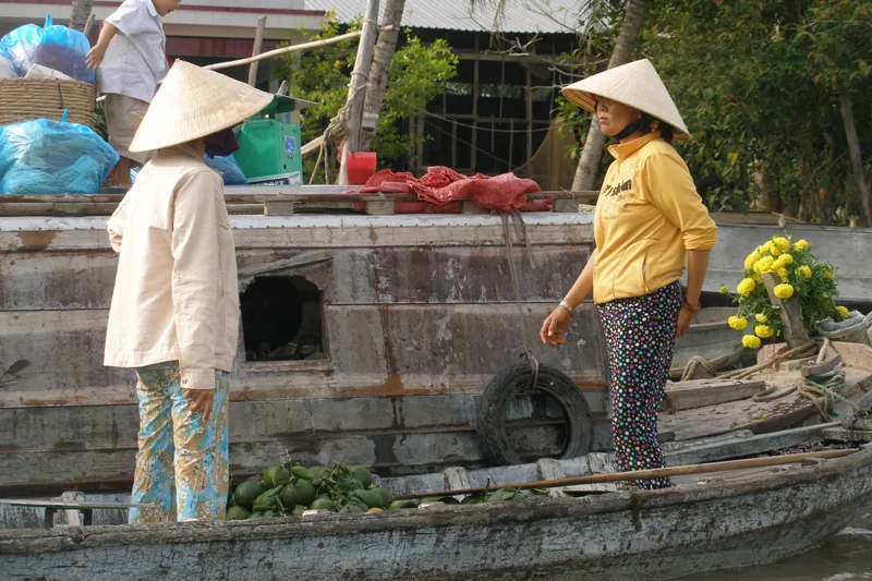 Two women in conical hats trade fruit between wooden boats at a Mekong Delta floating market.