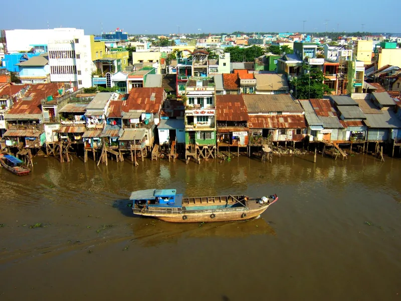 A wooden boat drifts past stilt houses lining a muddy river in My Tho, Mekong Delta.