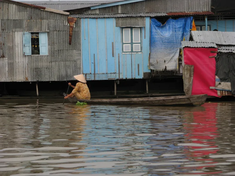 A woman in a conical hat rows a small wooden boat past colorful stilt houses on the Mekong Delta.