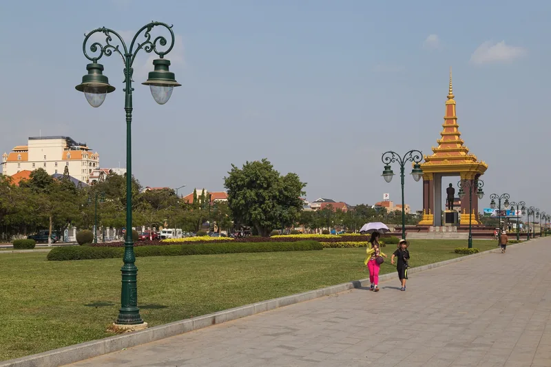 A golden-spired monument with a royal statue stands beside a riverside park promenade in Phnom Penh.