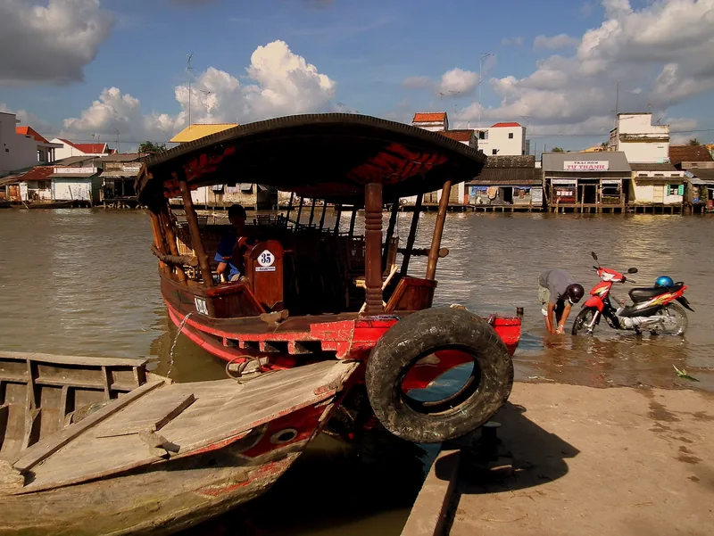 A wooden riverboat docks along a Mekong Delta riverbank while a man washes his motorbike in the shallow water.