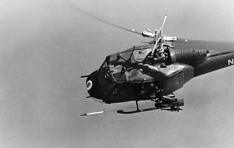 A vintage black-and-white photo shows a military helicopter armed with rockets flying against a plain sky.
