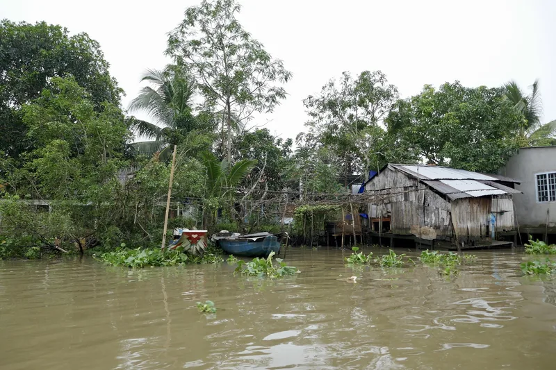 A wooden house on stilts and small boats line the muddy riverbank in the Mekong Delta.