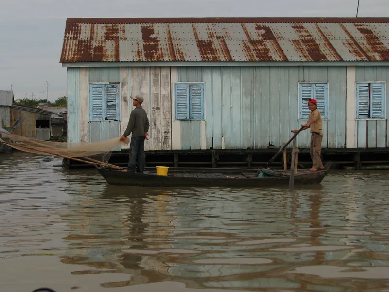 Two men fish from a wooden boat beside a stilted floating house on the Mekong Delta waterways.