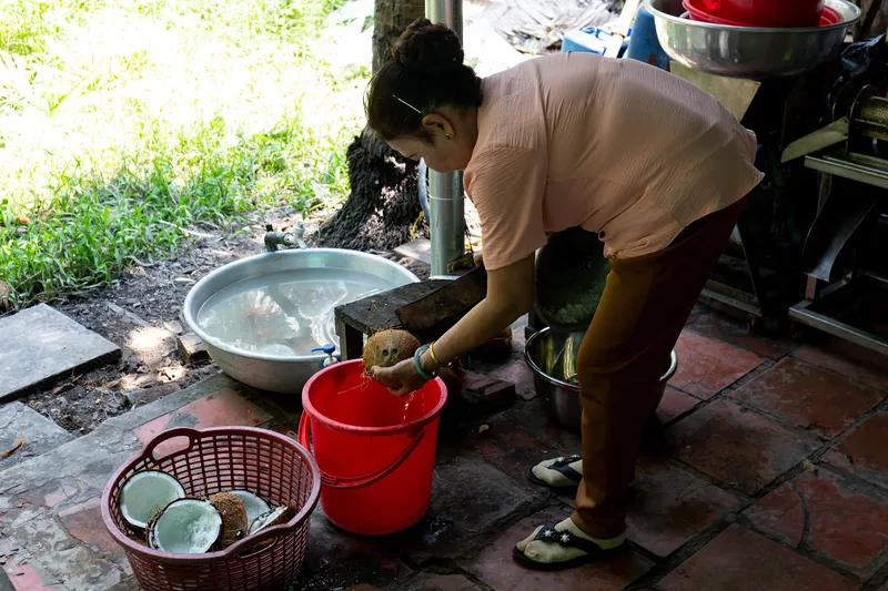 A woman washes coconut shells in a red bucket at an outdoor kitchen in the Mekong Delta.