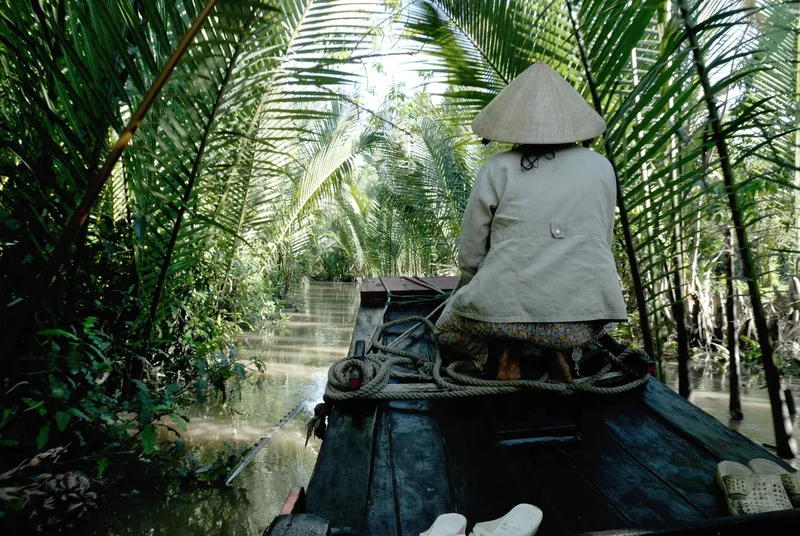 A conical-hat wearing rower sits at the bow of a wooden boat gliding through a narrow, palm-shaded Mekong Delta canal.