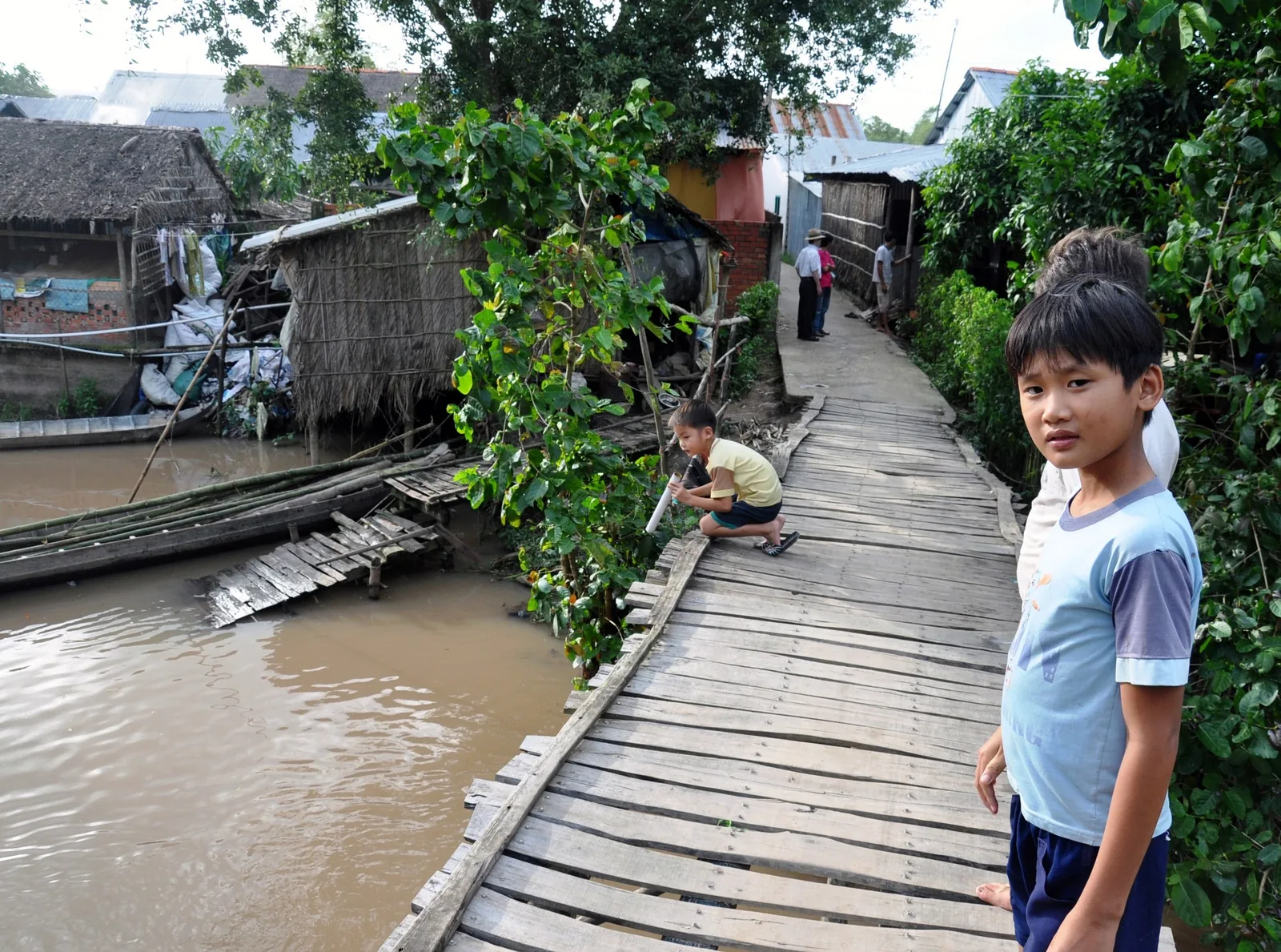 A boy stands on a wooden plank walkway beside a muddy canal lined with thatched houses in the Mekong Delta.