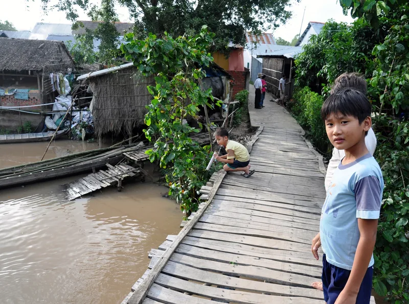 A boy stands on a wooden plank walkway beside a muddy canal lined with thatched houses in the Mekong Delta.