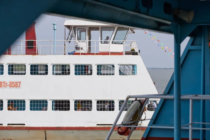 A white passenger ferry with a control cabin crosses the river in the Mekong Delta, framed by another boat's blue structure.