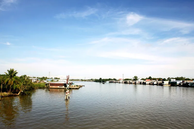 A wide river lined with fish farm rafts and floating houses under a bright sky in Ca Mau's Song Doc area.