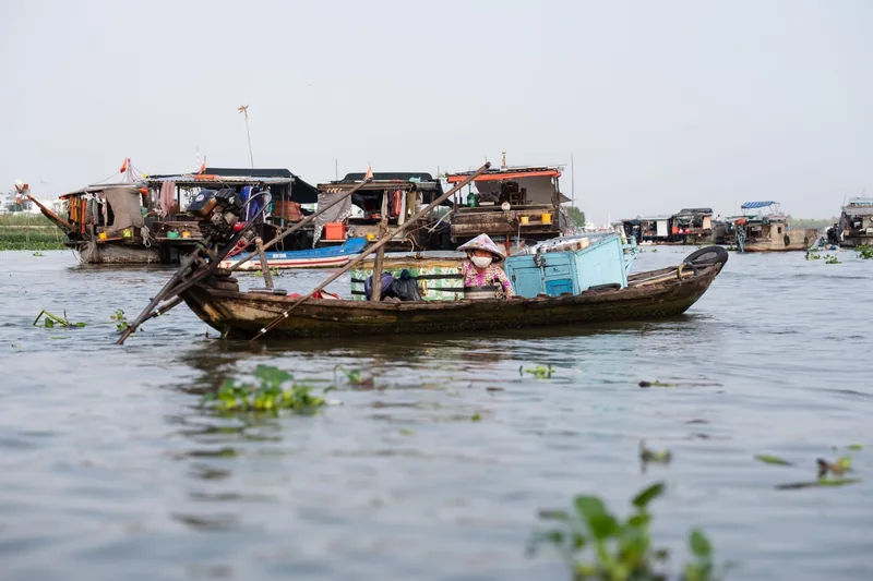A woman in a conical hat rows a small boat past floating houseboats on the Mekong Delta river.