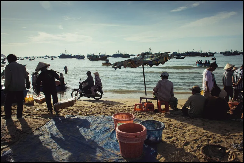 Fishermen gather on Mui Ne beach with baskets and a motorbike as boats crowd the water offshore.