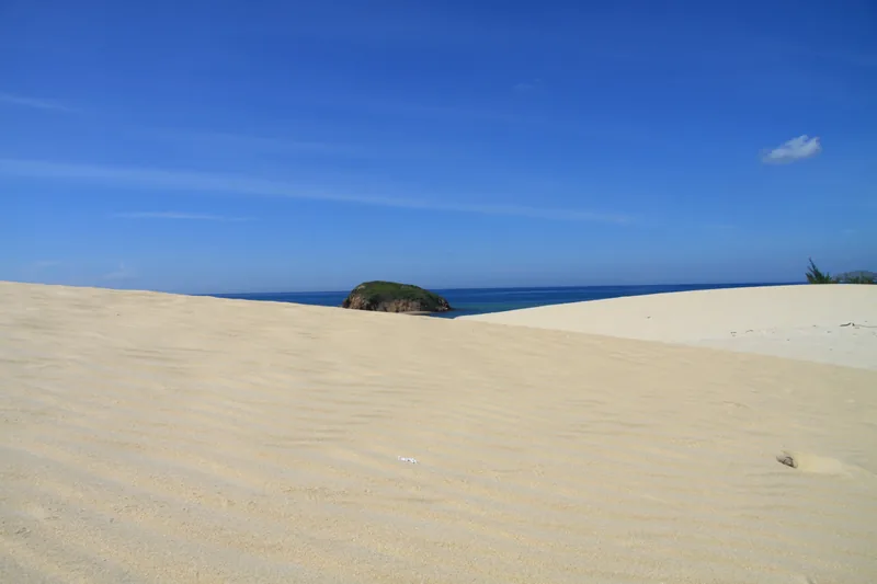 Wind-rippled sand dunes stretch toward the blue sea with a small green islet in Binh Thuan, Vietnam.