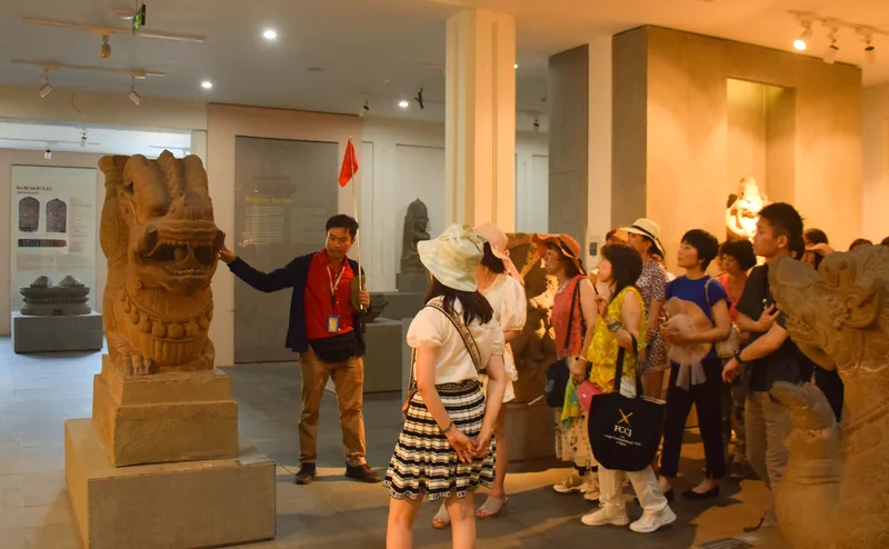 A tour guide with a flag leads visitors past sandstone lion sculptures inside a Da Nang museum gallery.