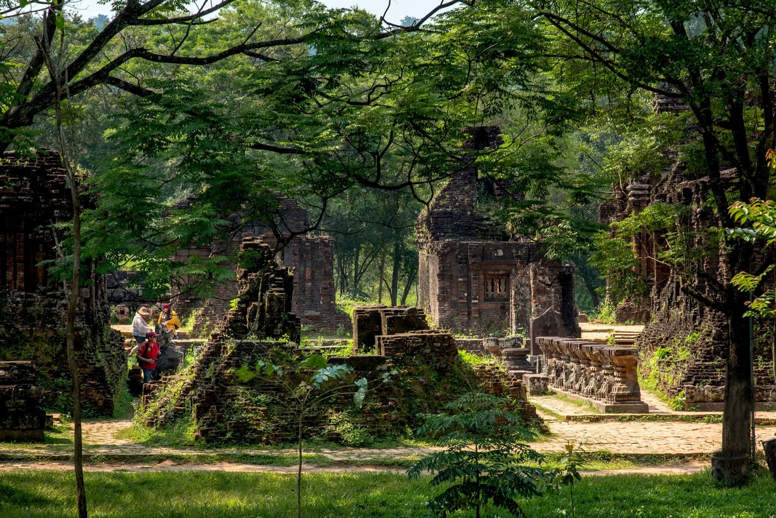 Ancient brick tower ruins at My Son sanctuary sit among lush trees with visitors strolling the pathways below.