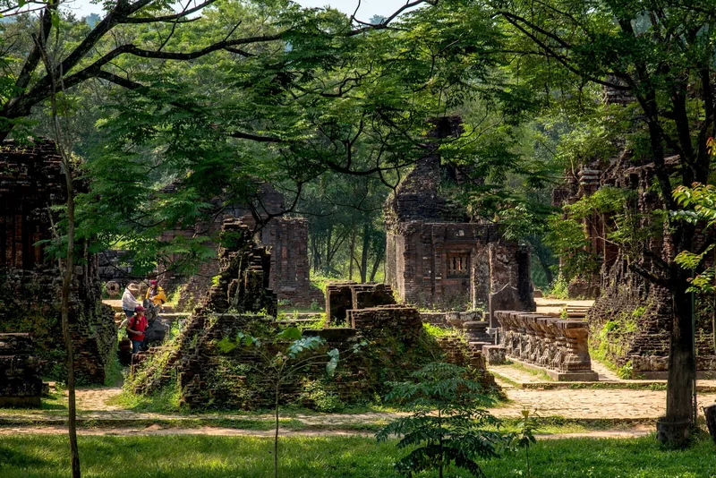 Ancient brick tower ruins at My Son sanctuary sit among lush trees with visitors strolling the pathways below.