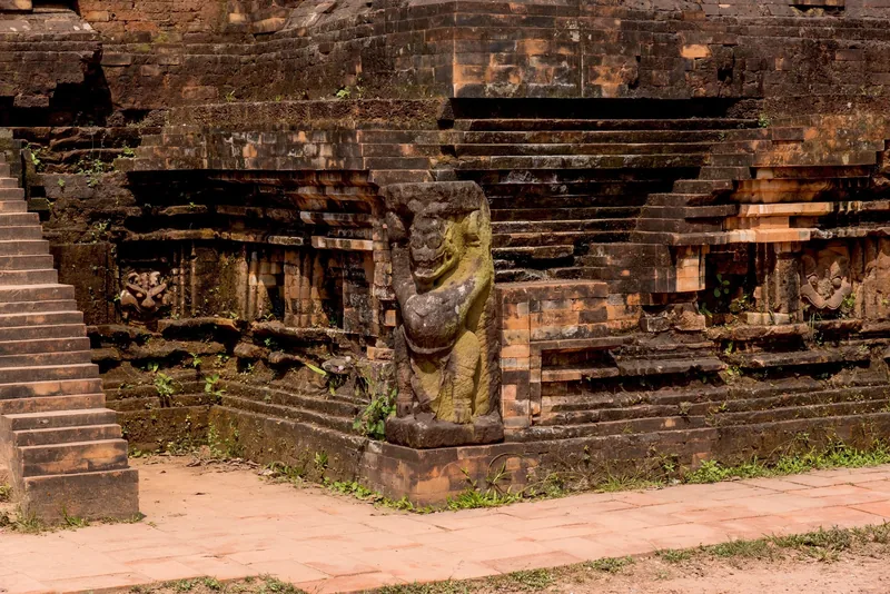 A weathered stone guardian statue stands at the base of an ancient brick tower at My Son sanctuary.