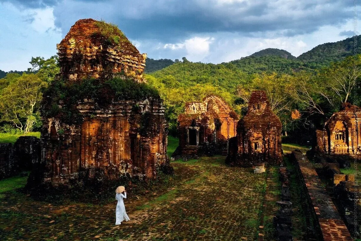 A woman in a white ao dai and conical hat walks among the ancient brick towers of My Son Sanctuary surrounded by green hills.