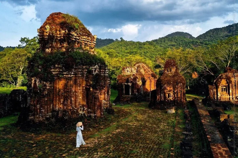 A woman in a white ao dai and conical hat walks among the ancient brick towers of My Son Sanctuary surrounded by green hills.