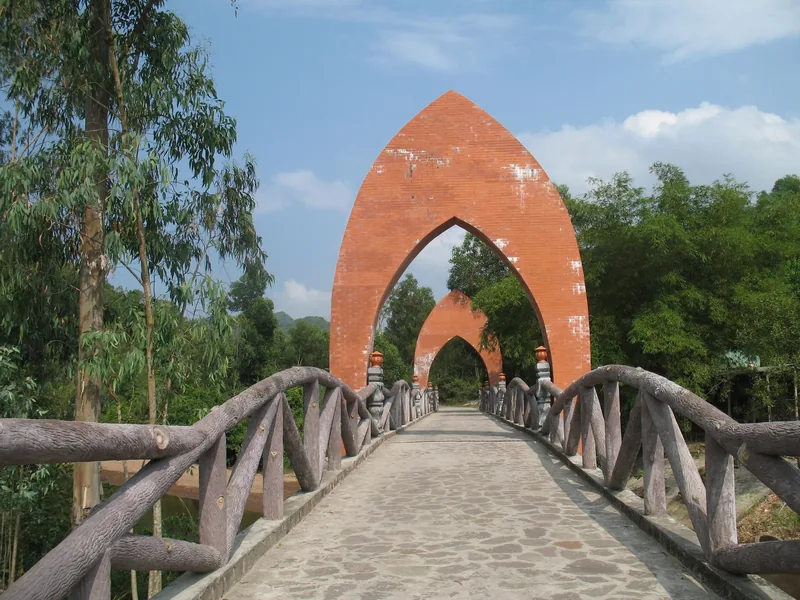A stone footbridge with tall pointed brick arches leads through greenery near My Son.