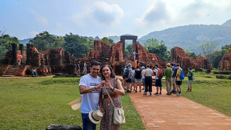 Tourists pose with a heart-shaped hand gesture before ancient brick ruins at My Son Sanctuary, surrounded by hills