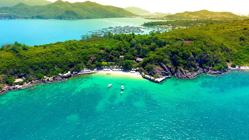Aerial view of a small sandy cove with turquoise water and anchored boats along the forested coastline of Nha Trang.