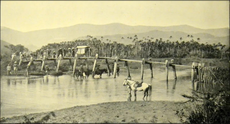 Historic sepia photograph of villagers crossing a wooden bridge over a river near Nha Trang, with a horse standing in the.