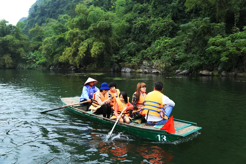 Tourists in life jackets ride a small wooden boat along a green river surrounded by limestone hills in Ninh Binh.