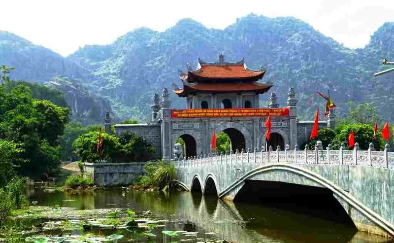 A stone bridge leads to the ornate gateway of Hoa Lu ancient capital, surrounded by limestone karst mountains in Ninh Binh.