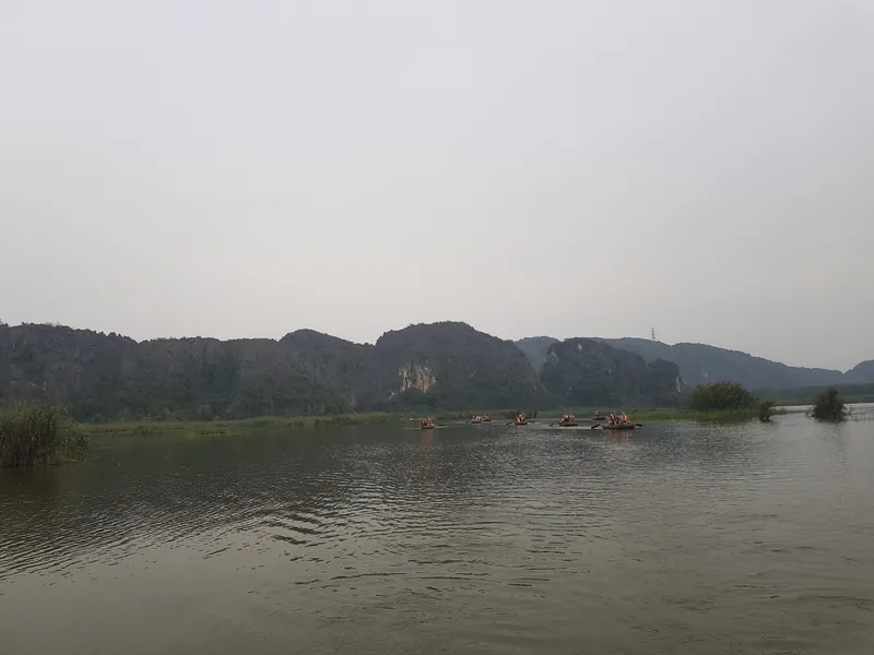 Small boats with orange-vested tourists glide across the calm river at Van Long Nature Reserve, Ninh Binh, framed by.
