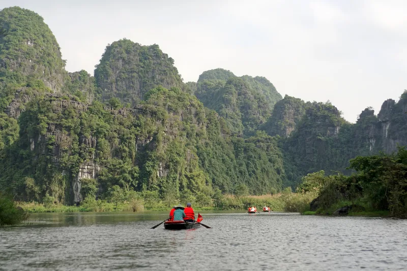 Rowboats with life-vested passengers glide along a river beneath towering limestone karsts in Trang An, Ninh Binh.