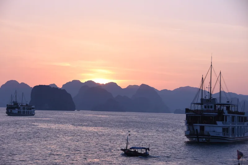 Cruise boats and a small vessel drift across Ha Long Bay as the sun sets behind limestone karsts.