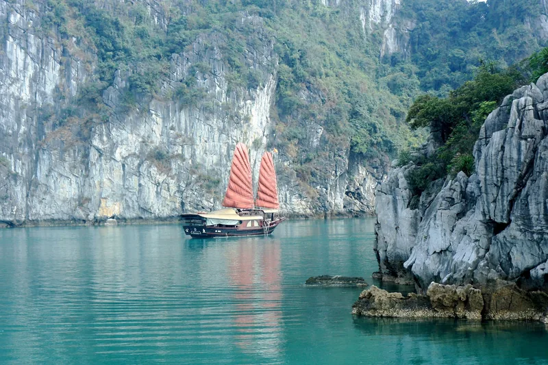 A red-sailed junk boat glides between towering limestone karsts in Ha Long Bay.